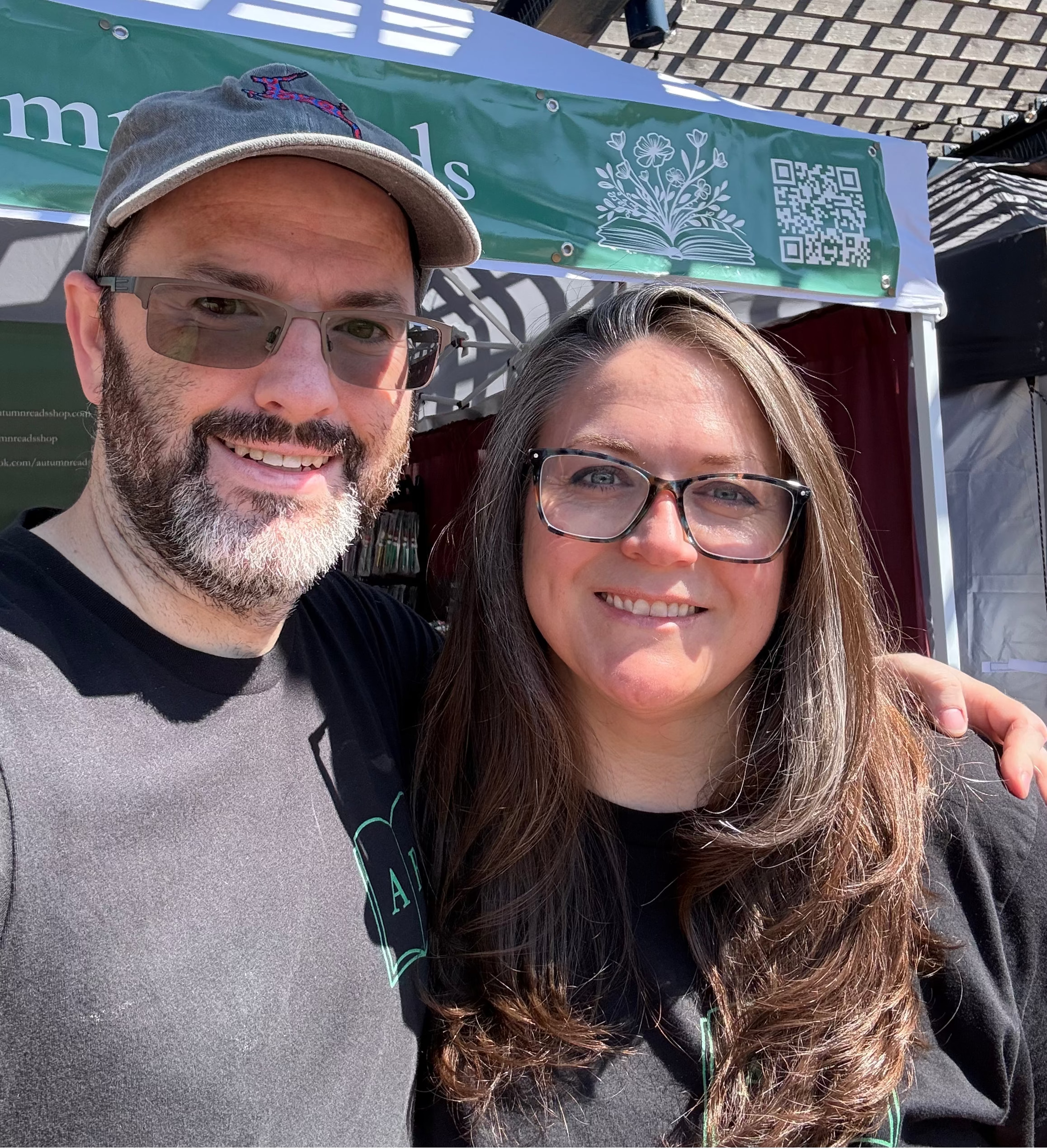Two people standing together with a green Autumn Reads LLC  banner and canopy in the background